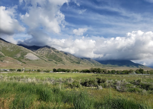 A scenic landscape featuring green fields, mountains, and a cloudy blue sky.